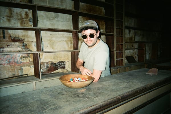 The writer, Alex, standing behind a counter with sunglasses on. A bowl of multicolored dice sits in front of him.